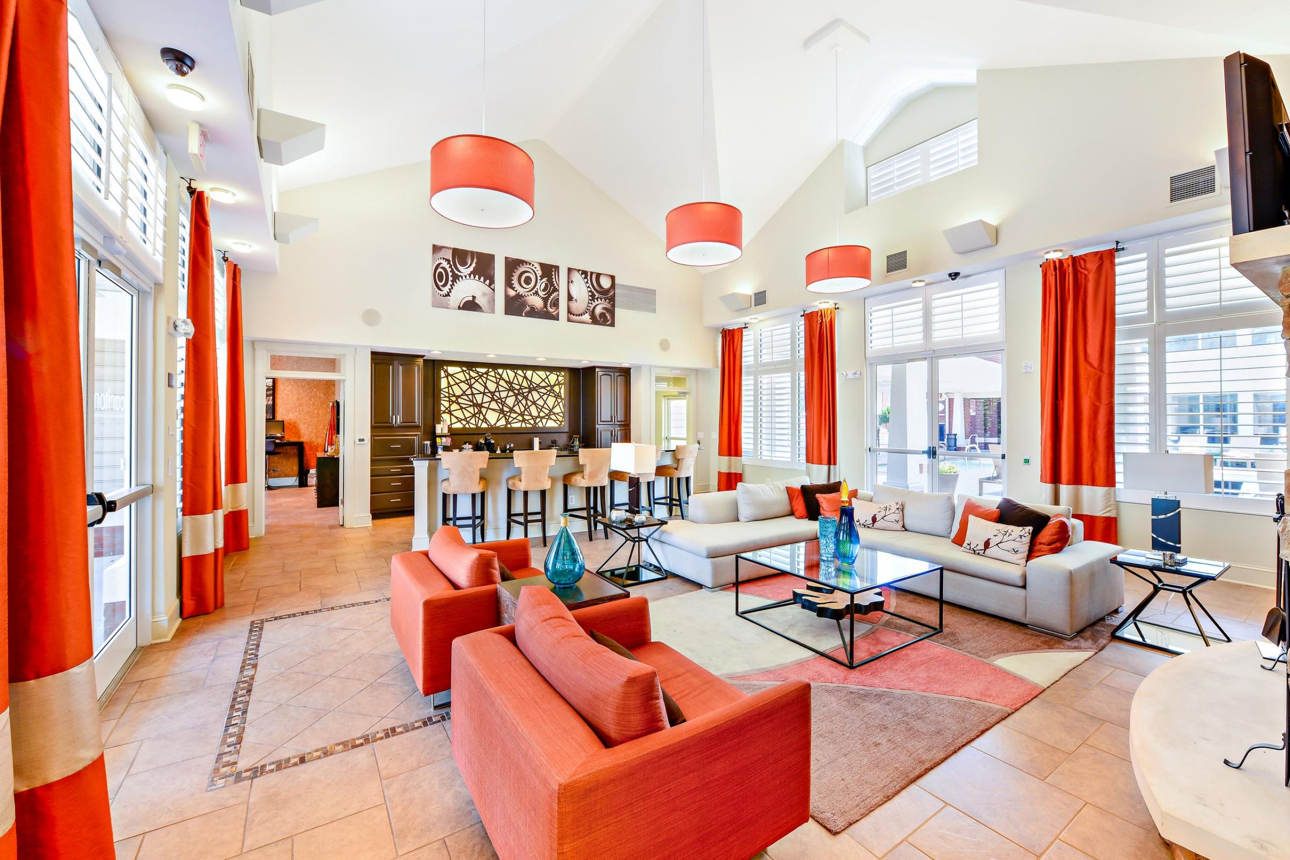 Modern lounge with red sofas, orange accents, bar seating, large windows, and bright lighting.