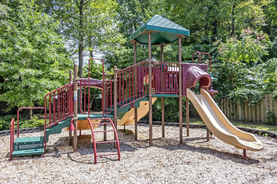 Colorful playground structure with slides and ladders on wood chips, surrounded by trees and greenery.
