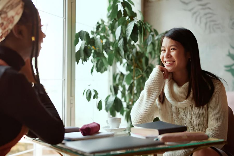 Two women sit at a table by a window, chatting and smiling, with notebooks and a coffee cup in front of them.