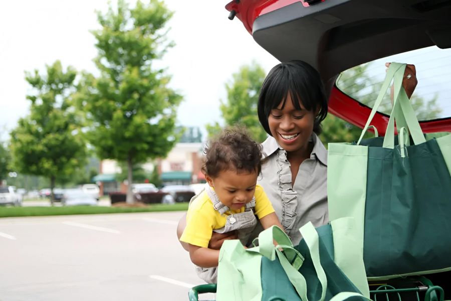 Woman and child unload groceries from the trunk of a car in a parking lot on a sunny day.
