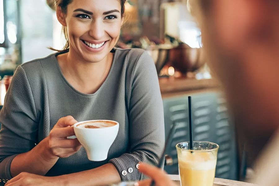 Smiling woman holding a coffee cup, sitting at a table in a café, talking to someone.
