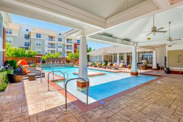 Covered patio with ceiling fans overlooks a pool and lounge chairs at an apartment complex on a sunny day.