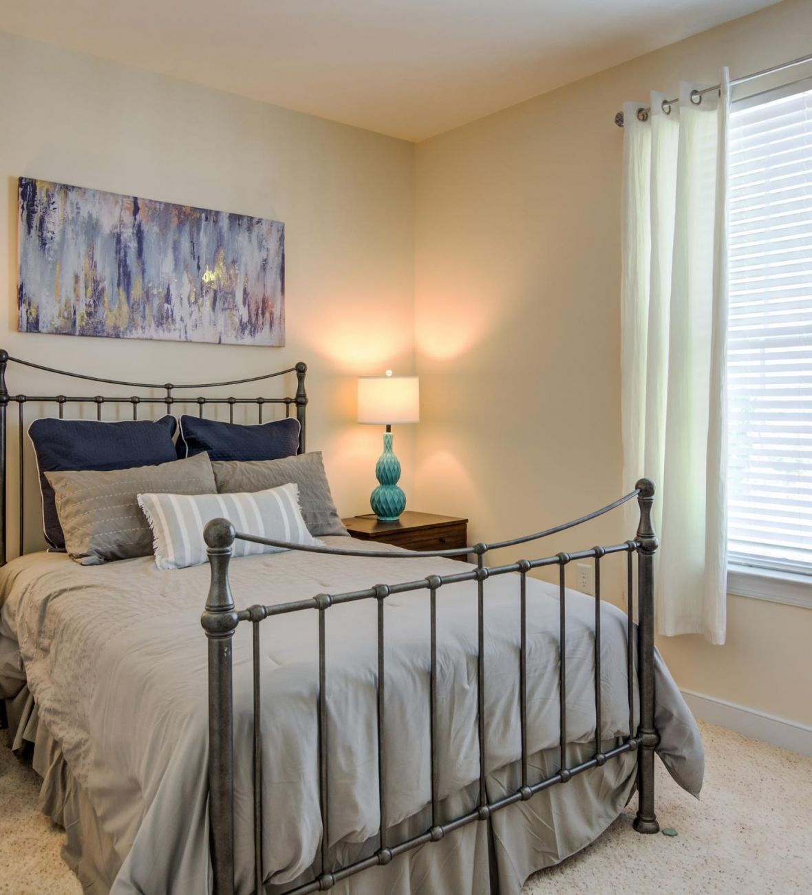 A cozy bedroom with a metal bed frame, gray bedding, two lamps, and a large window with white curtains.