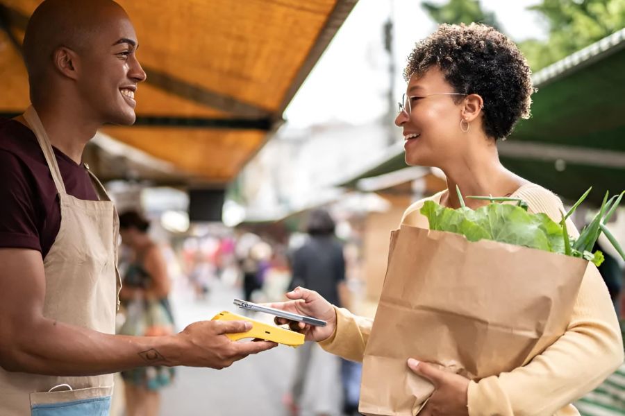 Smiling woman pays vendor with phone at an outdoor market, holding a paper bag of fresh vegetables.