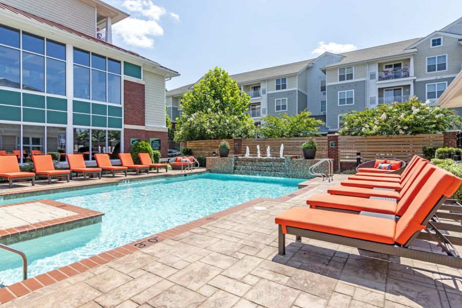 Outdoor swimming pool with orange lounge chairs, water fountains, and apartment buildings in the background.