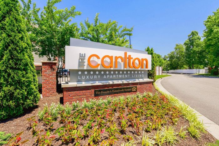 Sign for The Carlton Luxury Apartment Living, surrounded by plants and trees on a sunny day.