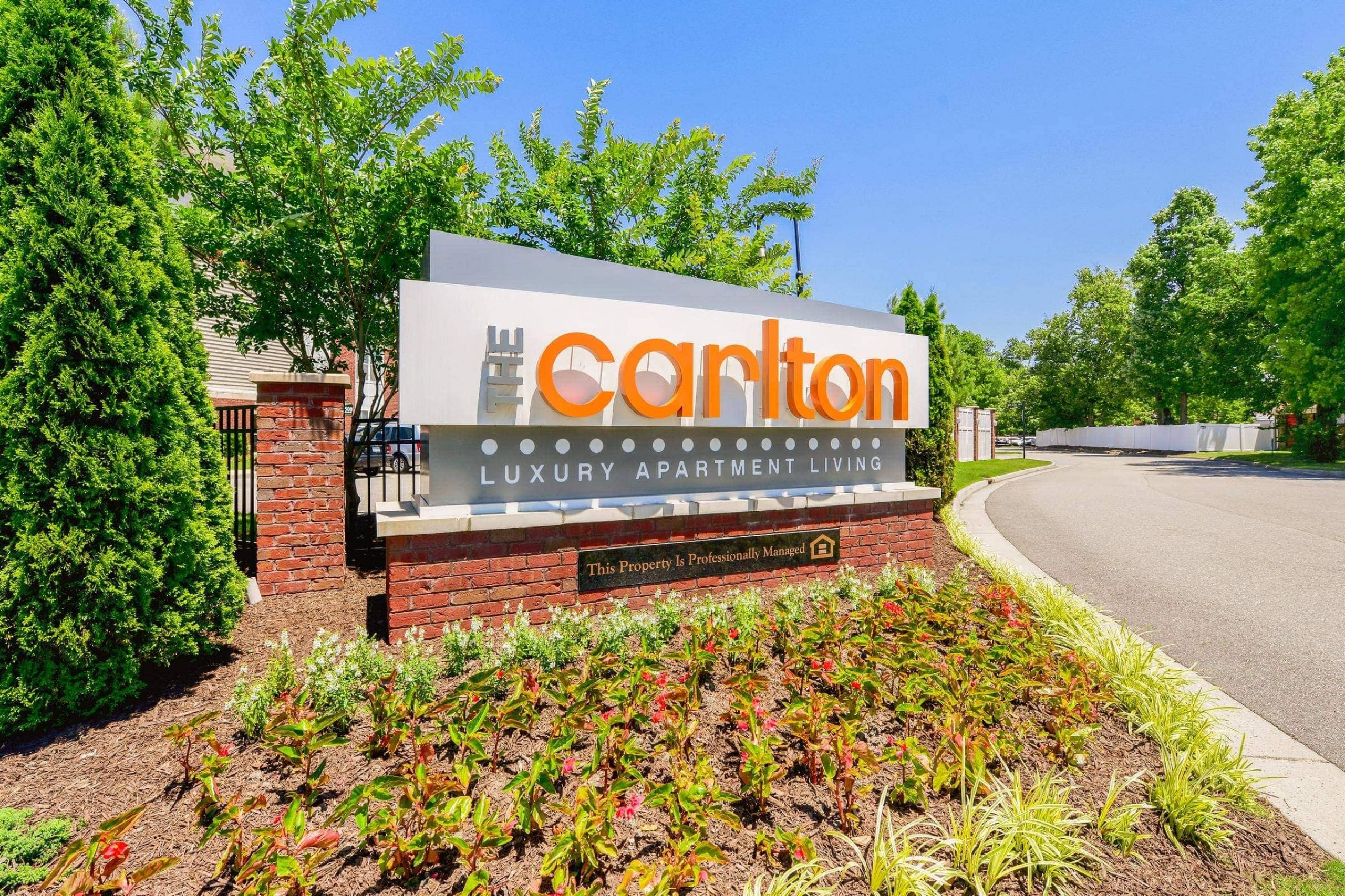 Sign for The Carlton Luxury Apartment Living, surrounded by plants and trees on a sunny day.