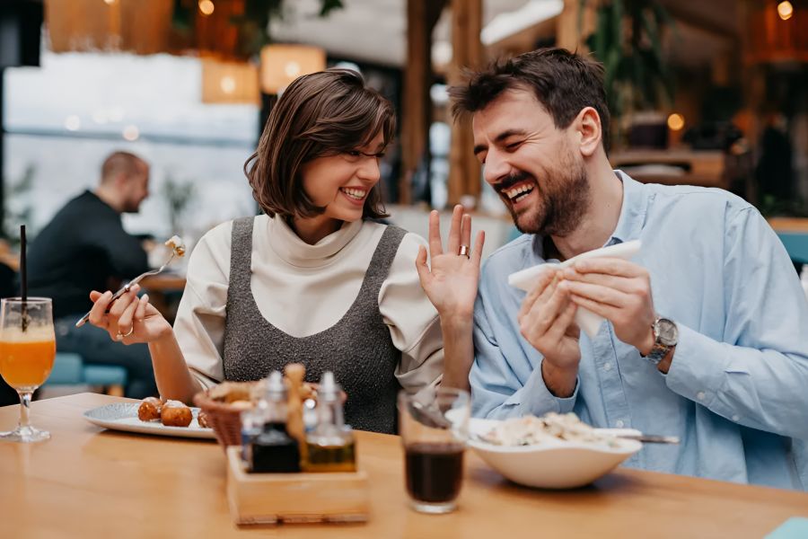A man and woman laugh together while eating at a restaurant, enjoying food and drinks.