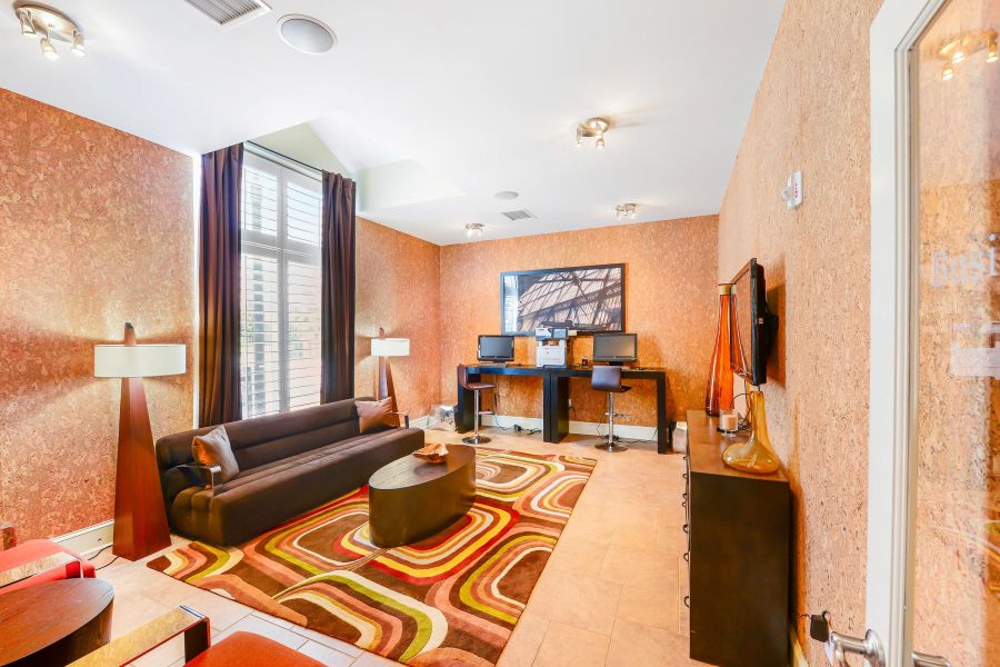 Modern lounge with brown sofa, three computers on a desk, abstract rug, and large windows with curtains.