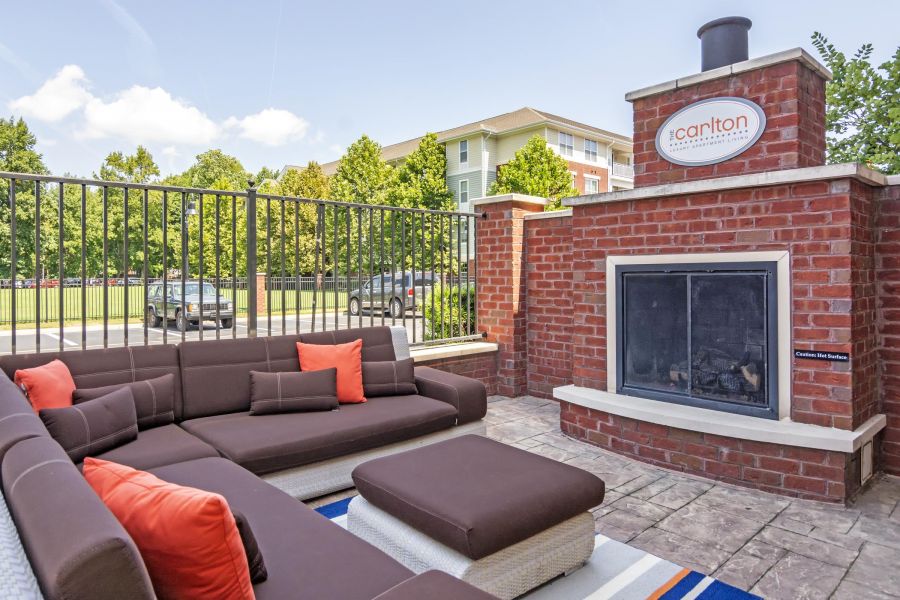 Outdoor patio with brown sectional sofa, orange pillows, and a brick fireplace near an apartment building.